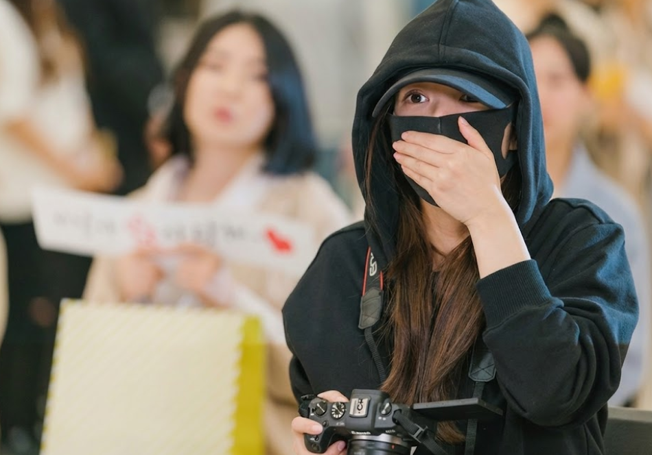Personagem Sung Deok-mi vestida com roupa preta e segurando câmera fotográfica profissional como fã de K-pop.
