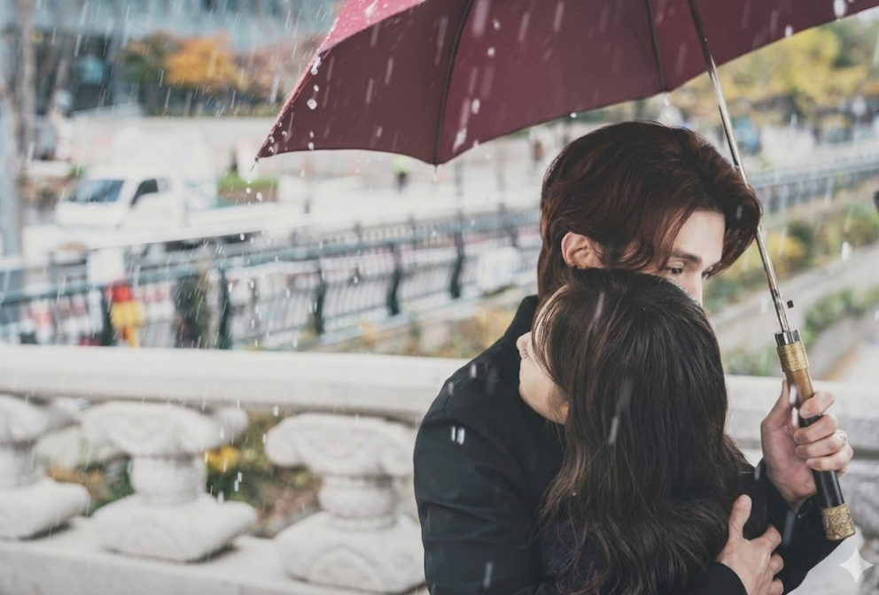 Lee Dong-wook segurando um guarda-chuva vermelho ao lado de Jo Bo-ah na chuva.