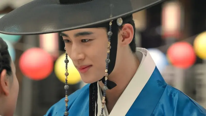 Do Joon sorrindo de forma galanteadora enquanto usa roupas nobres de época e um chapéu tradicional.