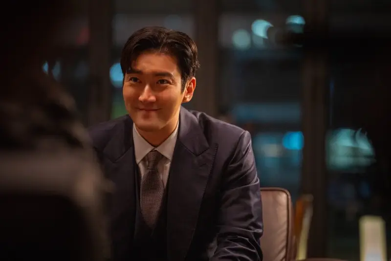 Choi Si Won como Hong Min Beom, sentado em uma poltrona de escritório, segurando um copo de whisky com olhar sombrio.