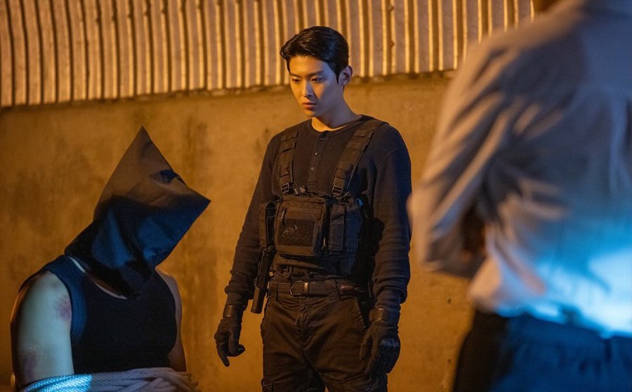 Dex com olhar desafiador e uniforme de agente especial em ambiente de operação noturna.