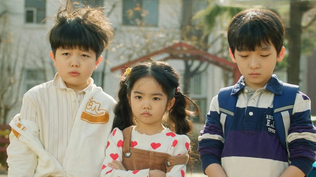 Cena do dorama Family by Choice: Oh Eun-seo sorrindo ao lado de dois meninos no parque, representando a forte amizade de infância.