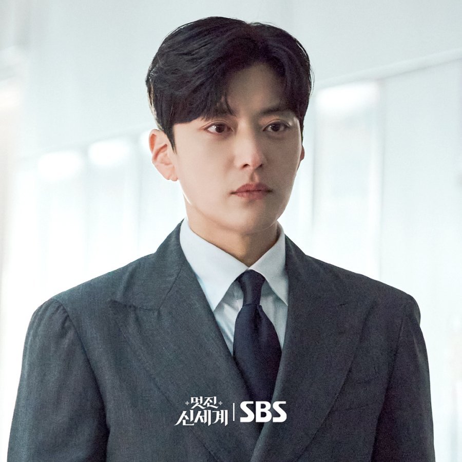 Jang Seung Jo com expressão serena e sorriso discreto, vestindo terno claro, em um ambiente luxuoso com iluminação baixa.