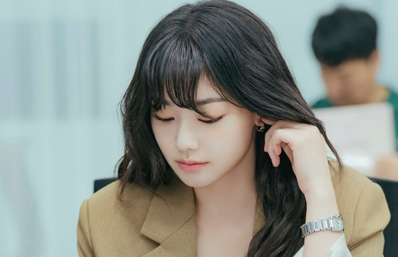 A atriz Kang Mi Na sorrindo em um ambiente corporativo moderno.