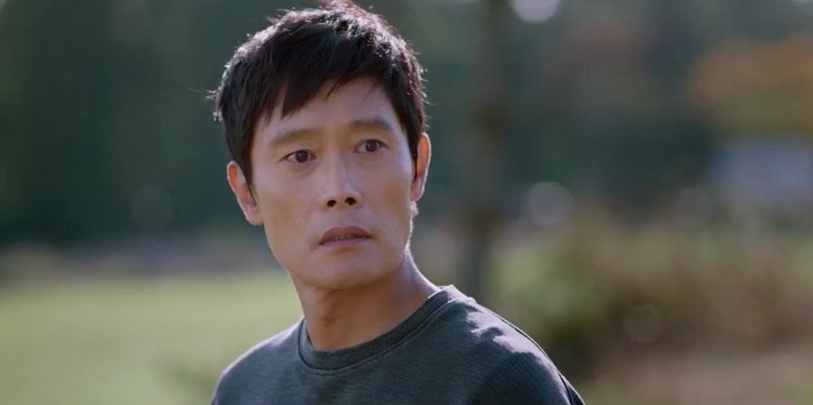 Lee Byung-hun como um comerciante de meia-idade, sentado em um banco de madeira em frente a uma loja simples, olhando para o mar.