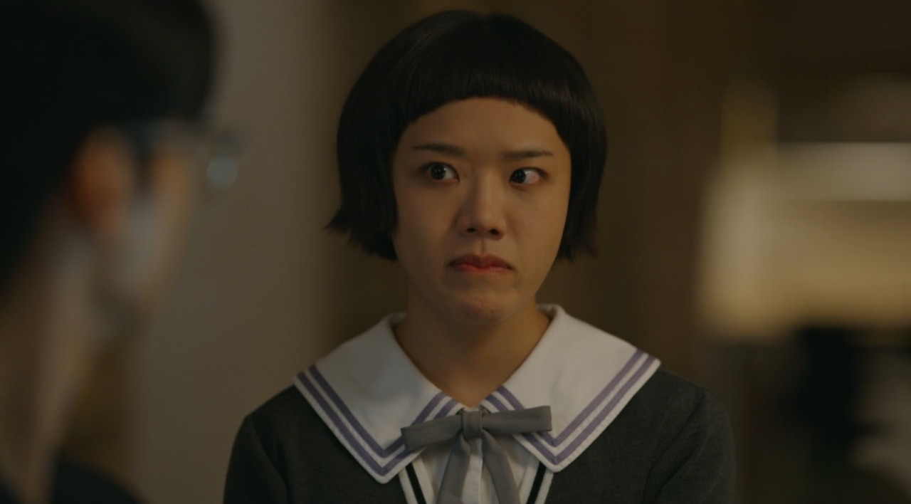 Kim Hyang Gi com óculos sem lentes, caderno na mão e expressão travessa, em um cenário escolar colorido.