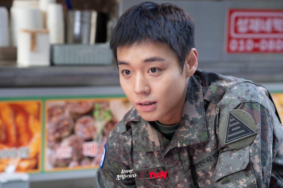 Jovem soldado com uniforme militar segurando uma frigideira e uma colher de pau, parado em uma cozinha industrial com outros soldados ao fundo.
