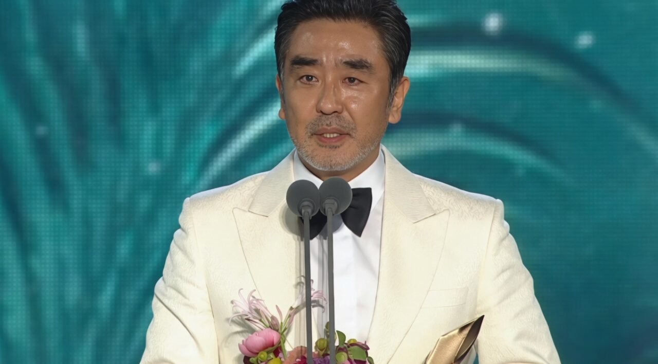 Ator de meia-idade em um palco de premiação, segurando um troféu dourado, vestindo terno escuro e sorrindo para a plateia.