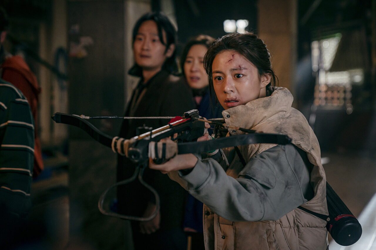 A atriz Go Youn Jung com um arco e flecha em cenário apocalíptico.