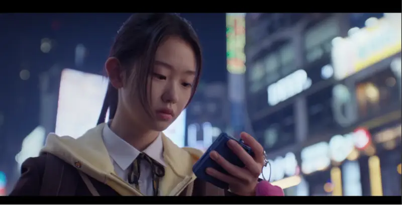 Min-ji visualizando a tela do celular instantes antes de ser atraída para a emboscada que resulta em seu desaparecimento