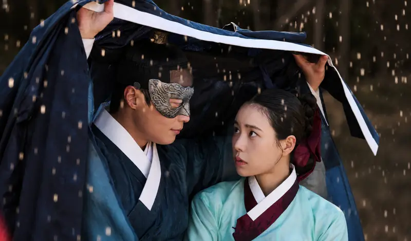 Ator sul-coreano cobrindo uma atriz com uma capa preta durante uma forte chuva.
