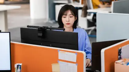 Funcionária Cha Ji Yoon, com expressão de cansaço diante da tela de um computador no ambiente de escritório.