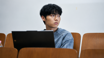 O personagem Lee Kang com uma expressão serena e observadora, contrastando com o ambiente acadêmico ao fundo em cena de Notas da Última Fila