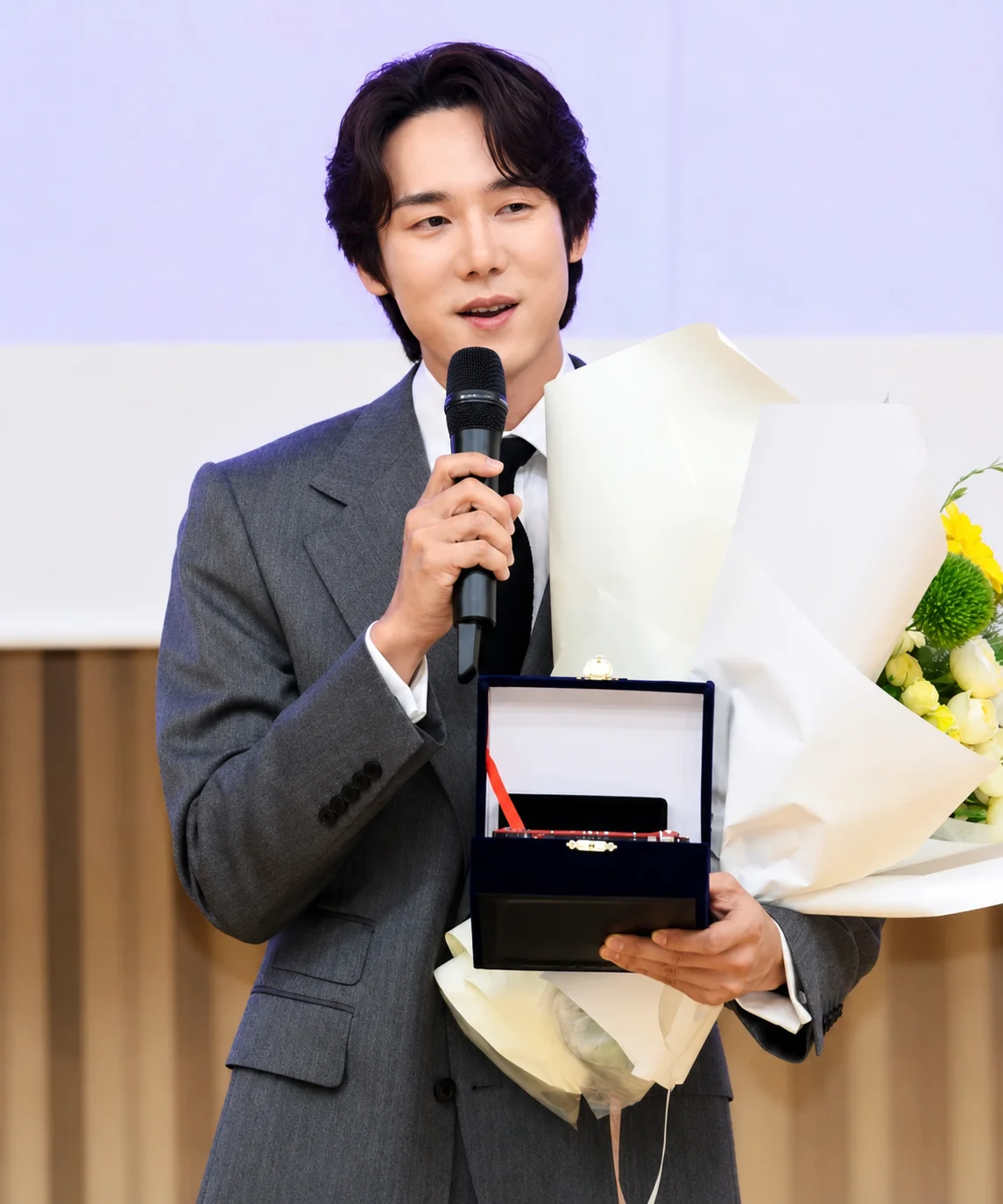 O ator Yoo Yeon Seok discursando no palco da premiação da SBS, com tom descontraído enquanto segura seu prêmio
