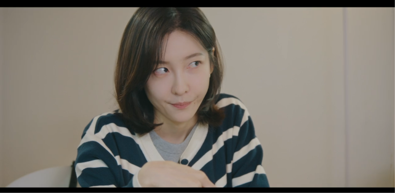 Personagem Cha Ji-yoon com expressão preocupada sentada em sua mesa de trabalho.