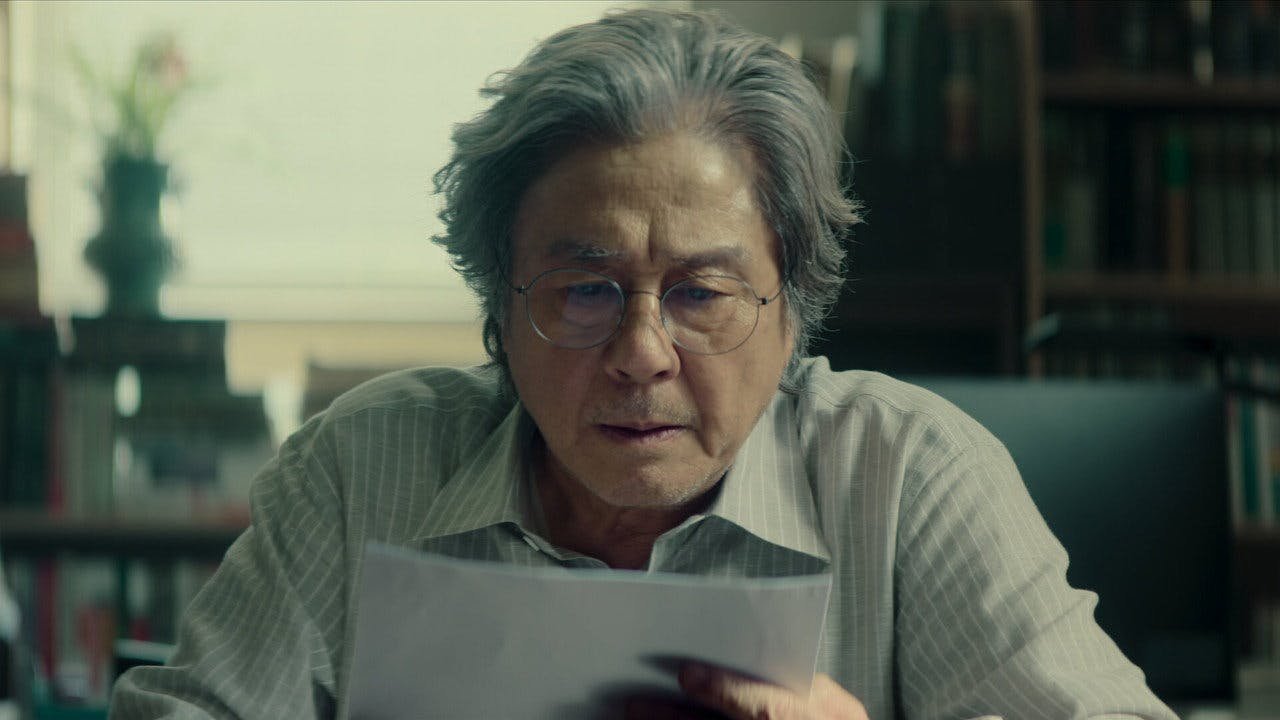 O professor Heo Mun Oh analisando o texto do aluno em seu escritório, demonstrando confusão e obsessão pela narrativa.